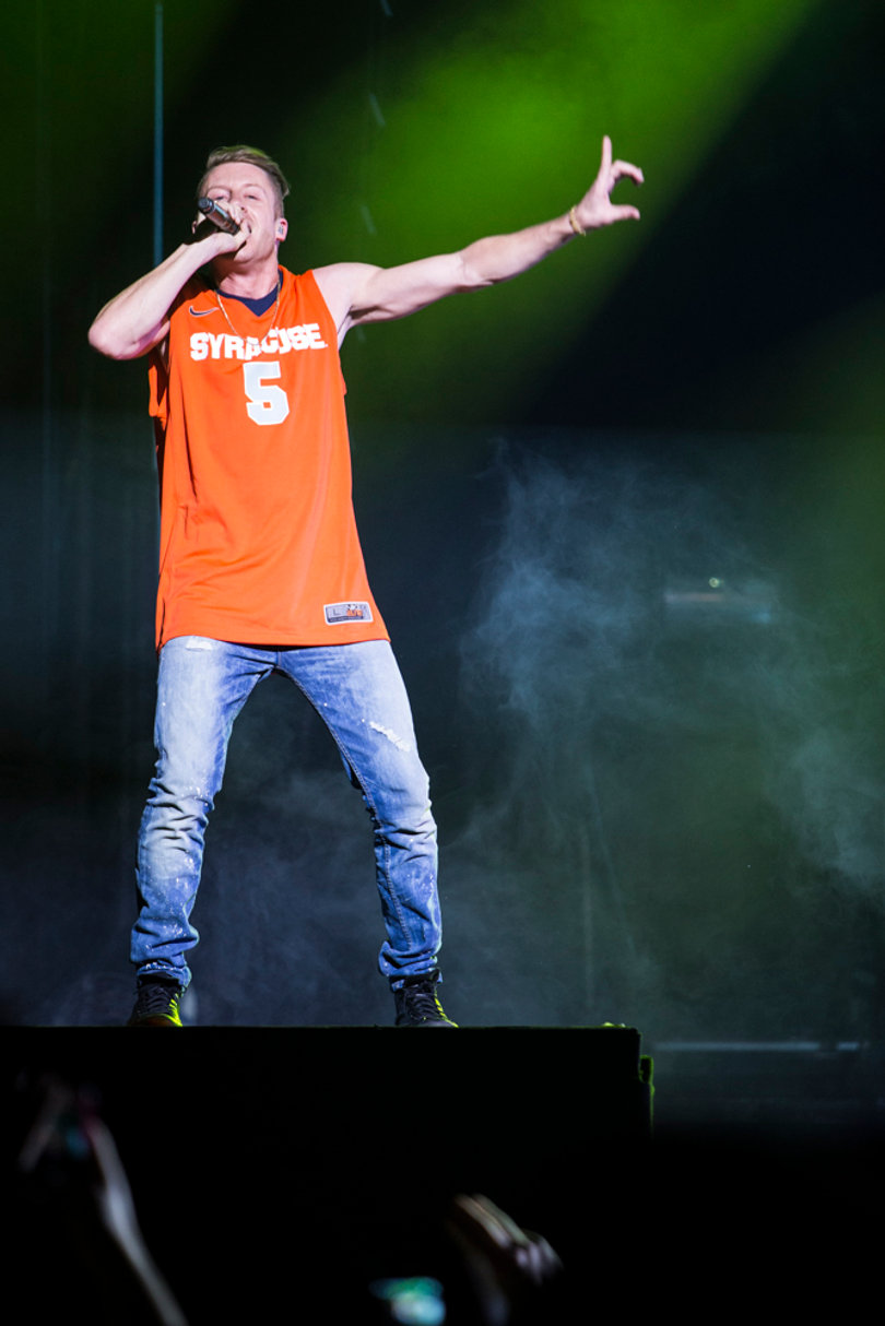 Macklemore stands atop a riser and raps along to his hit single "Thrift Shop," wearing a Syracuse jersey emblazoned with his last name on the back.