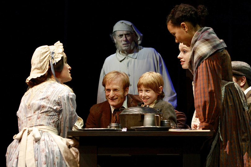 Tiny Tim and Bob Cratchit gather at the table to enjoy Christmas dinner with others while Scrooge and the ghost of Christmas present observe from the background.