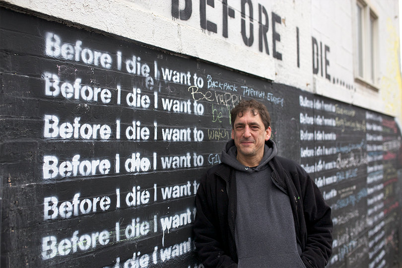 Charlie Sam, a local artist, stands by the Before I Die wall on South Geddes street which he helped paint.