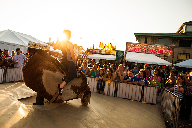 Jordan Thomas, 16, of Orange County hangs on tight while trying to beat eight seconds on a mechanical bull in front of the stand Wine Slushies at The Great New York State Fair. Onlookers smile and hold other fair refreshments as they cheer on Thomas.