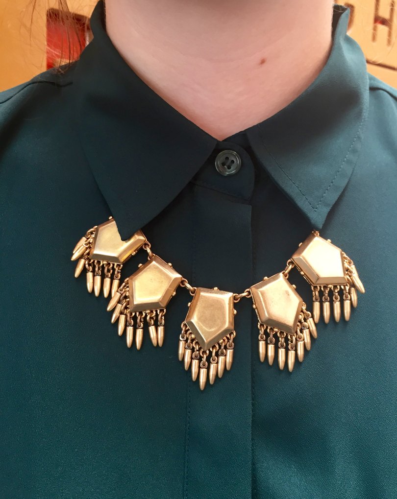 Anello rocks a chunky statement necklace that contrasts with her collared green shirt.