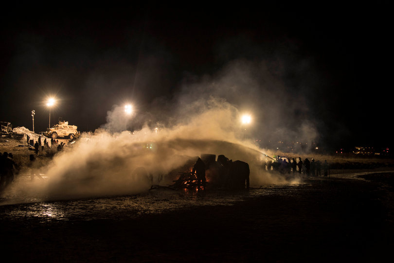Water protectors ignite fires for warmth for hypothermic victims of water cannons and as a symbol of sacred fire and tradition. They use blue tarps to protect the fires from the law enforcement water hoses.