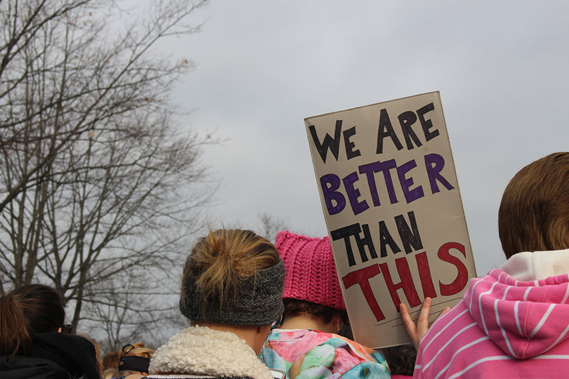 A woman in a pink "p*ssyhat" holds a sign that reads "we are better than this."