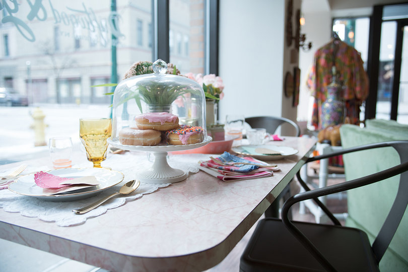 On one side of the room is a table with flowers, gold table settings and fresh pink glazed donuts covered by a glass cake dome.