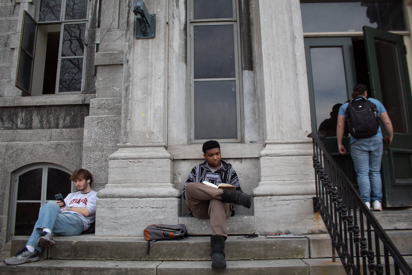 Two students sit outside the Hall of Languages reading and on their phone, while somebody else is walking into the building.