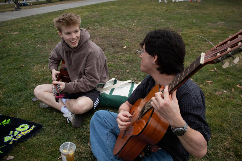 Two students sit in the grass and play guitar together.