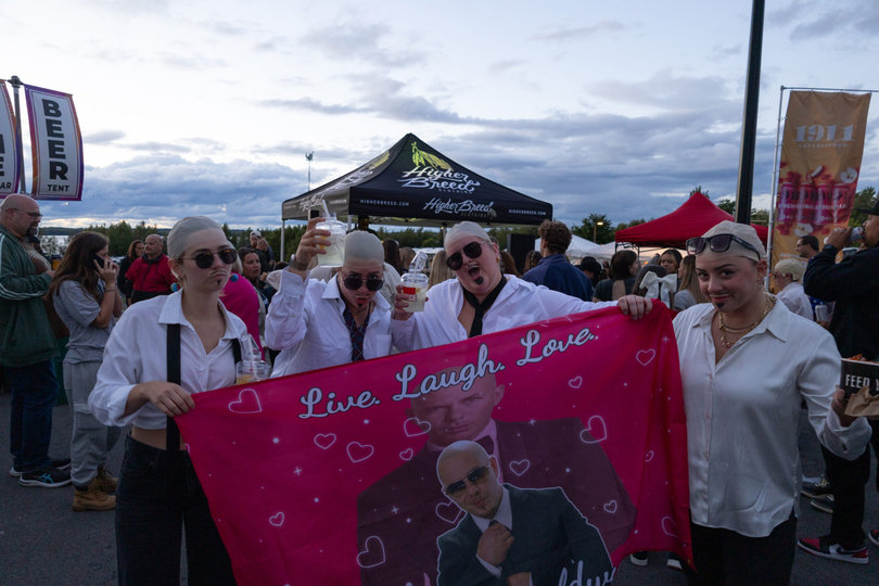 Costumed concertgoers dressed as Mr. Worldwide hold a flag emblazoned with a slogan of their love for Pitbull. These four attendees were inspired by a TikTok trend to wear the costumes. 

