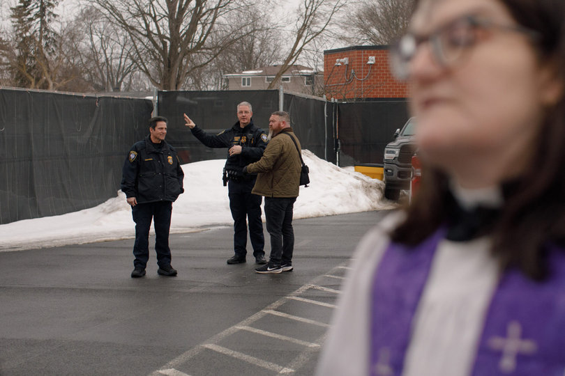 Two ICE agents stand talking to a man; the reverend is off to the side in the frame.