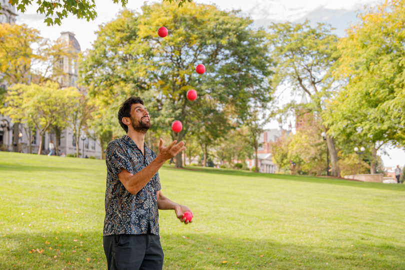 Josef Komissar juggles several red balls outside Syracuse University's Hall of Languages. Komissar, a mathematics Ph.D. student at SU, enjoys learning and teaching and about the math behind juggling.